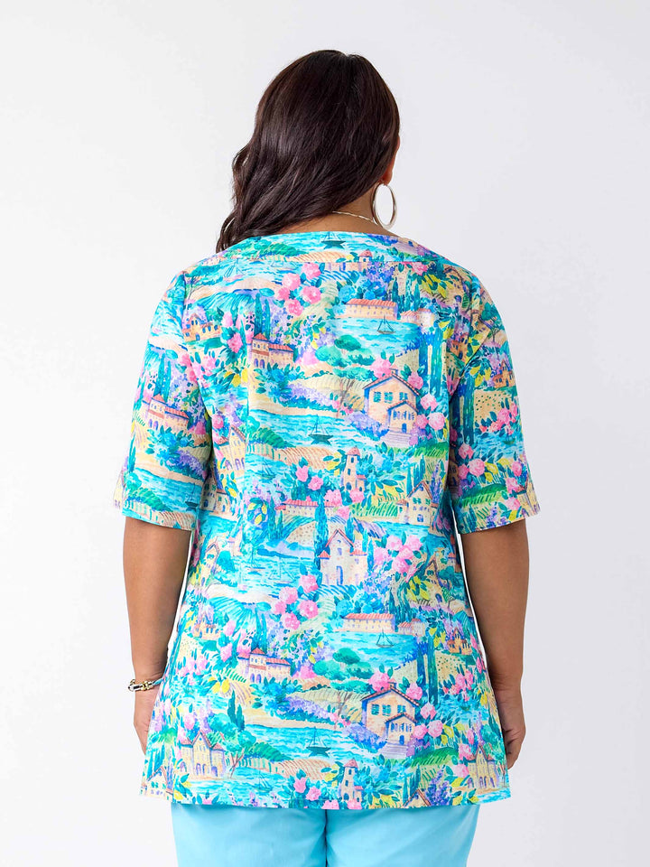 A woman shown from the back wearing a short-sleeve tunic top with a colorful, detailed landscape print featuring blue skies, green trees, pink flowers, and houses.