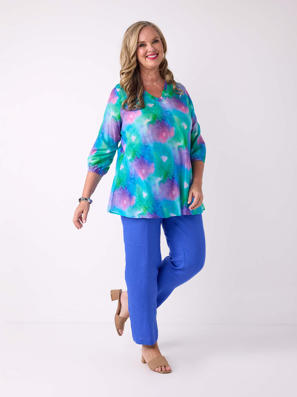 A woman wearing a v-neck silk cotton watercolour top in blue, green, and pink, with ruffle detail around the neck, paired with a cobalt blue linen pant