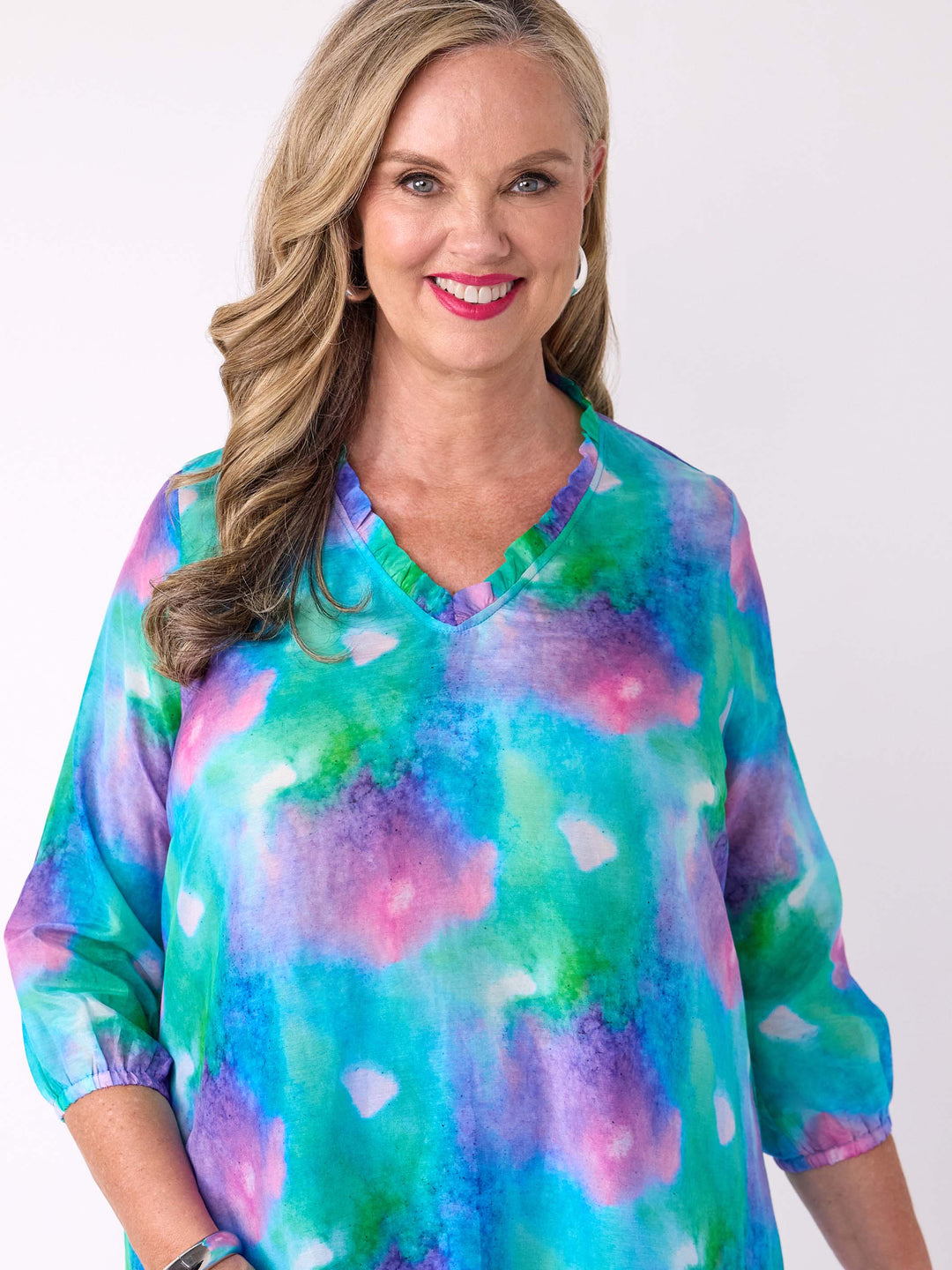 A woman wearing a v-neck silk cotton watercolour top in blue, green, and pink, with ruffle detail around the neck