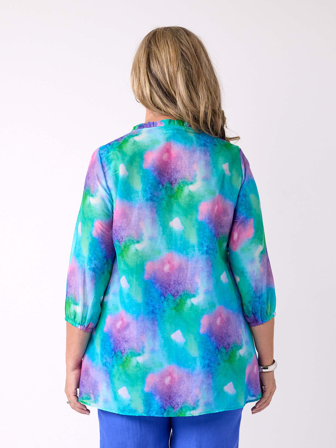 A woman viewed from the back wearing a v-neck silk cotton watercolour top in blue, green, and pink, with ruffle detail around the neck, paired with a cobalt blue linen pant