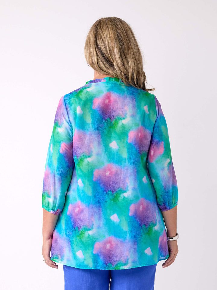 A woman viewed from the back wearing a v-neck silk cotton watercolour top in blue, green, and pink, with ruffle detail around the neck, paired with a cobalt blue linen pant