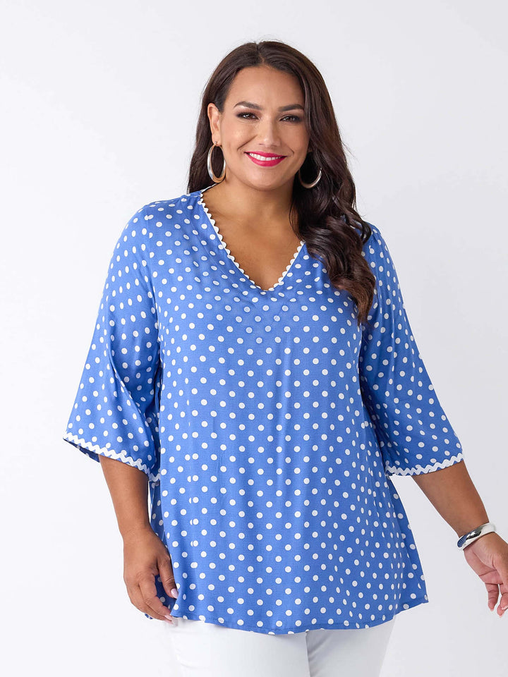 A woman wearing a blue top with white polka dots and ric rac trim details around the v-neckline and three-quarter sleeves
