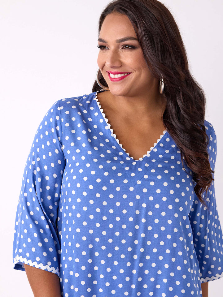 A woman wearing a blue top with white polka dots and ric rac trim details around the v-neckline and three-quarter sleeves