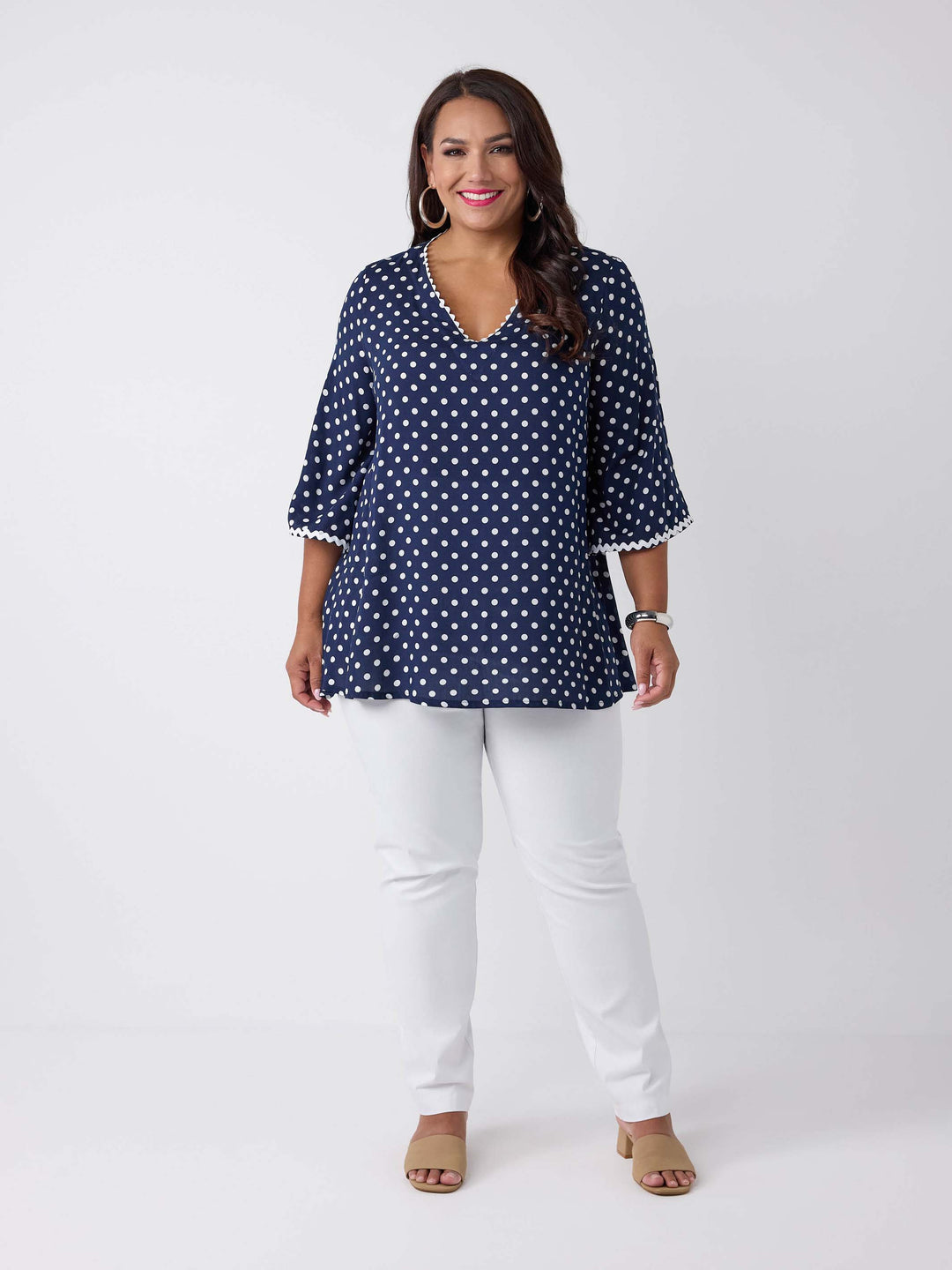 A woman wearing a navy blue V-neck top with white polka dots and ric rac trim on three-quarter sleeves and around neckline, paired with white pants