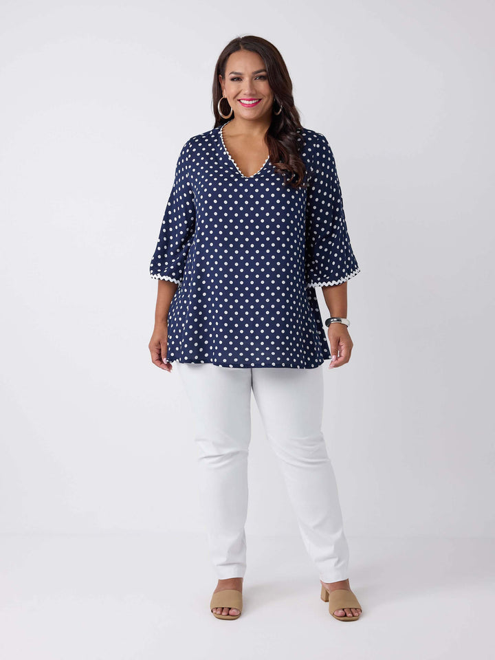 A woman wearing a navy blue V-neck top with white polka dots and ric rac trim on three-quarter sleeves and around neckline, paired with white pants