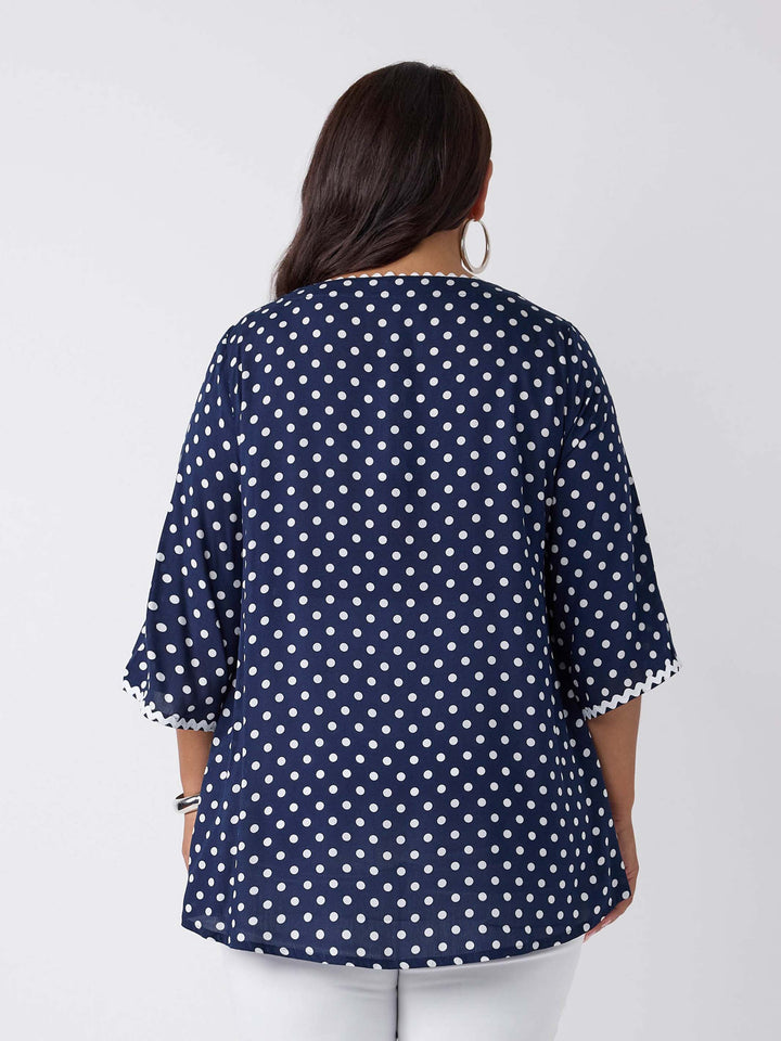 A woman wearing a navy blue V-neck top with white polka dots and ric rac trim on three-quarter sleeves and around neckline, paired with white pants