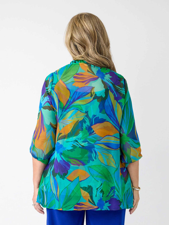 A woman viewed from the back wearing a vibrant, multicoloured abstract print georgette top with blue, green, orange, and purple tones, paired with bright blue pants