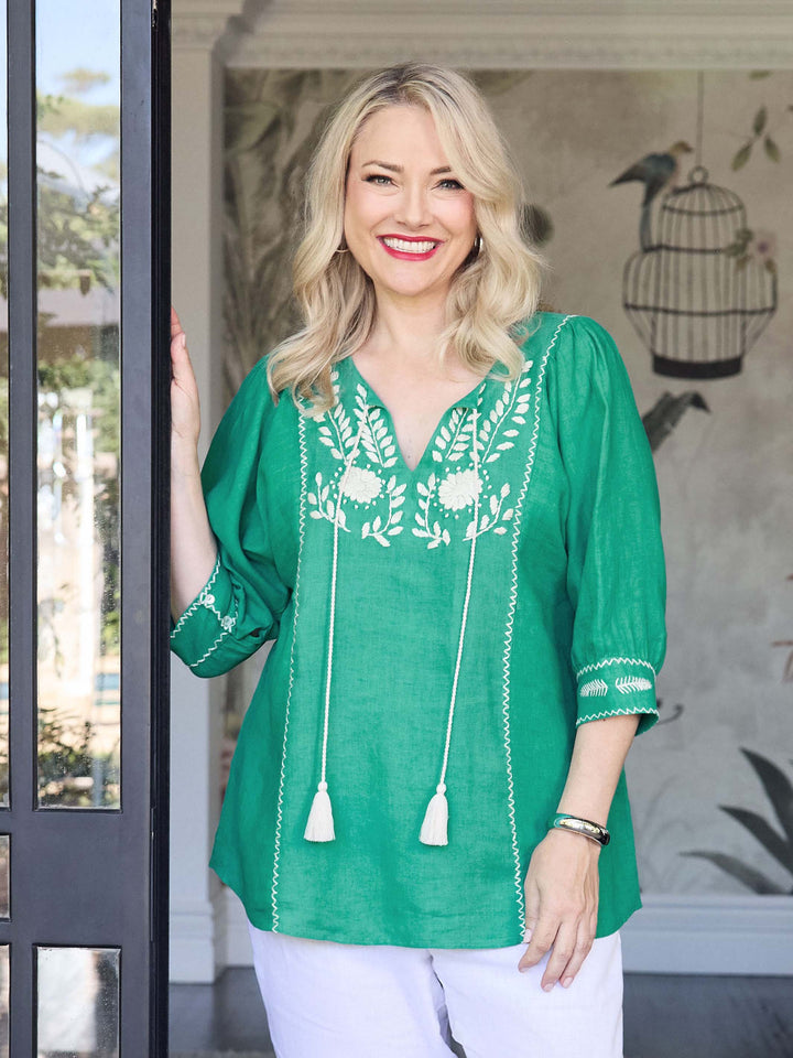 A woman wearing a vibrant emerald green embroidered linen top with white tassels and white pants