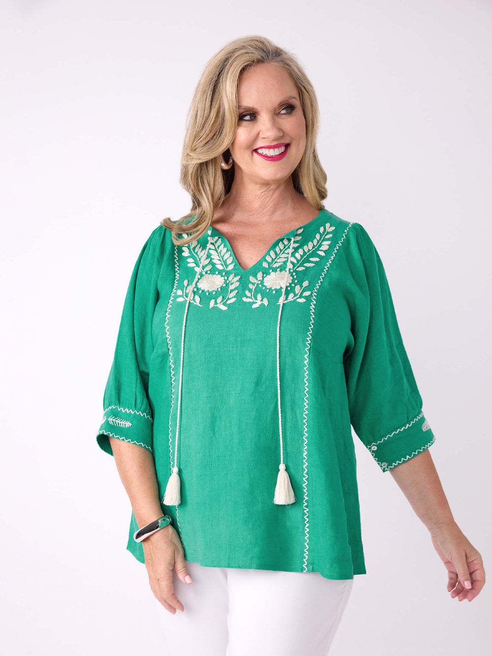 A woman wearing a bright emerald green short-sleeve top with white floral embroidery, drawstrings, and tassel details, paired with white pants 