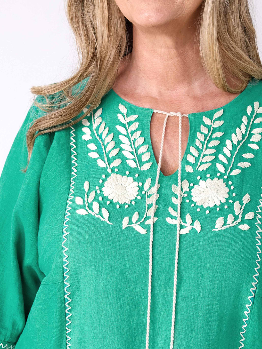 A close-up of a woman wearing a bright emerald green linen top with white floral and leaf embroidery around a v-neckline and front tie closure.