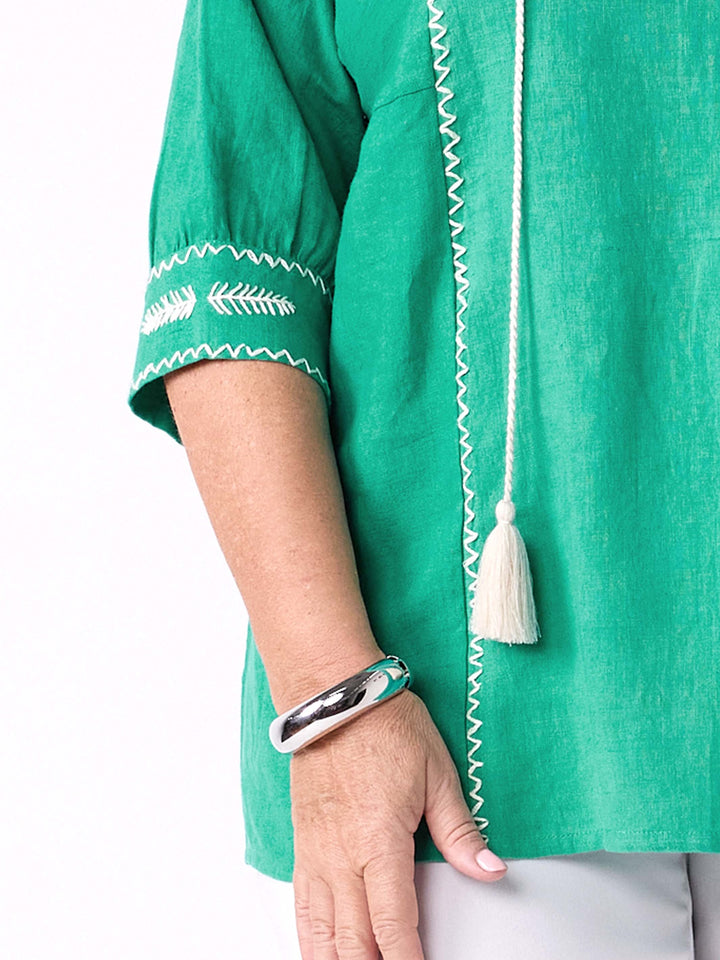 A close-up of a person wearing a bright emerald green linen top with white embroidered details on the sleeve and a vertical white cord with a tassel.