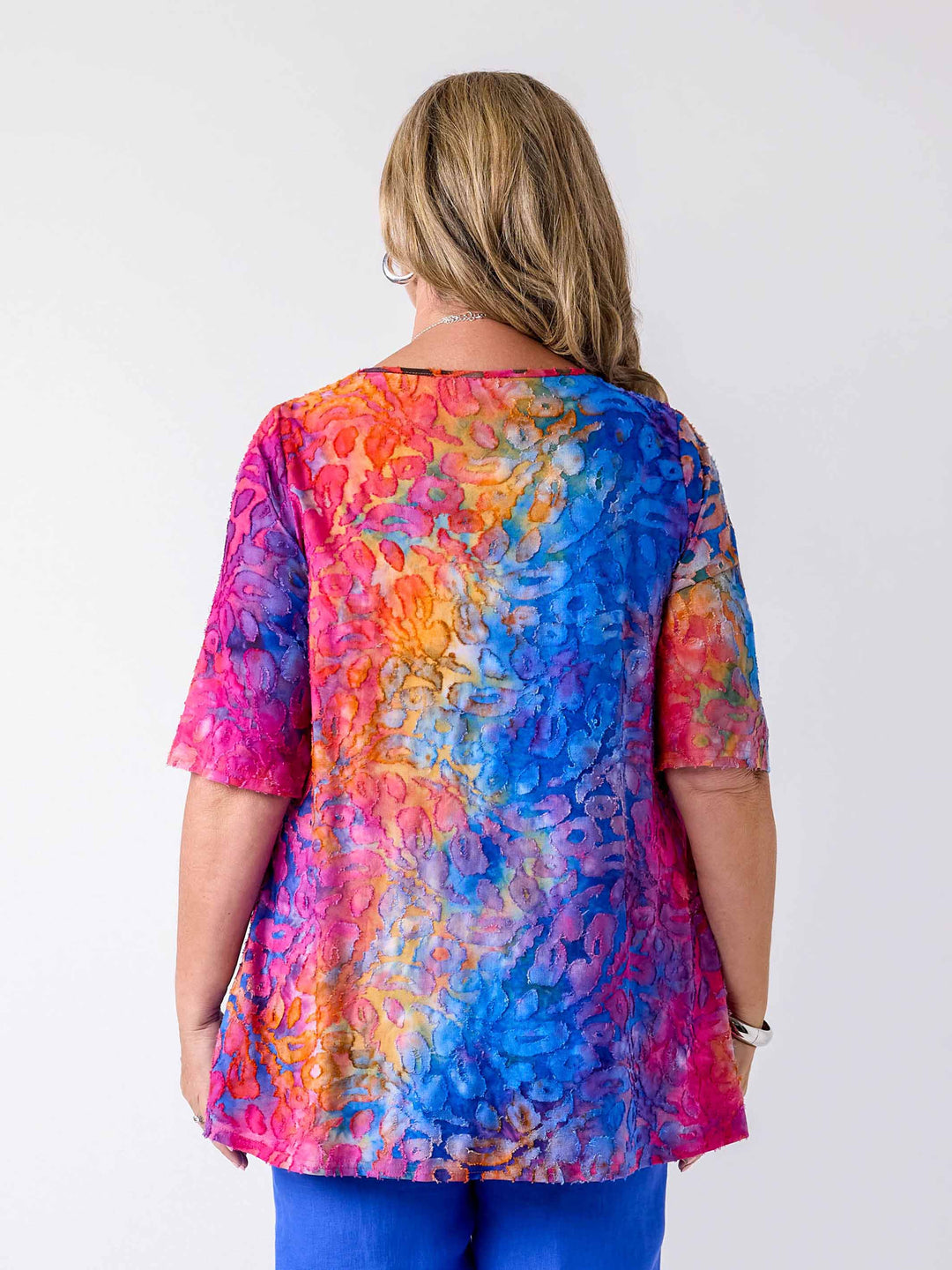 back view of woman wearing a colourful swing jersey top with blue and pink tone print with burnout detail, paired with blue pants