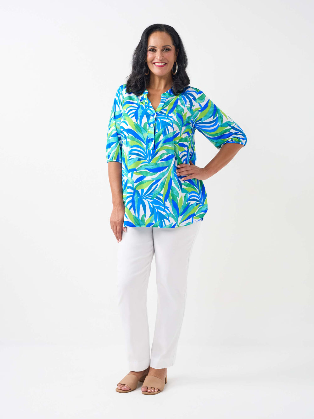 A woman wearing a vibrant tropical leaf print button-up shirt with three-quarter sleeves, paired with white pants