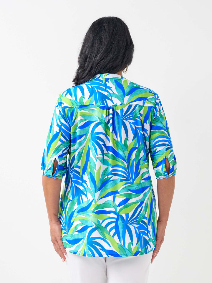 A woman viewed from the back wearing a short-sleeve button-up shirt with a bold tropical leaf print in blue, green, and white, paired with white pants