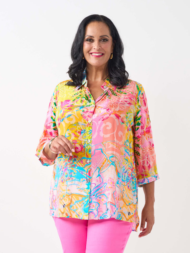 A woman waring a vibrant silky collared shirt with button front and colourful print in pink and yellow tone, paired with bright pink jeans