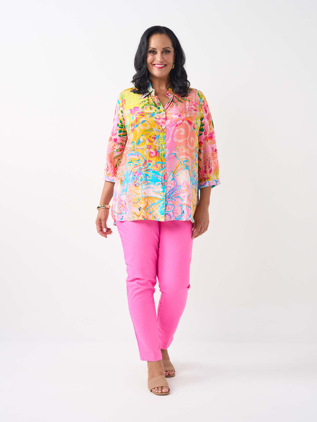 A woman waring a vibrant silky collared shirt with button front and colourful print in pink and yellow tone, paired with bright pink jeans