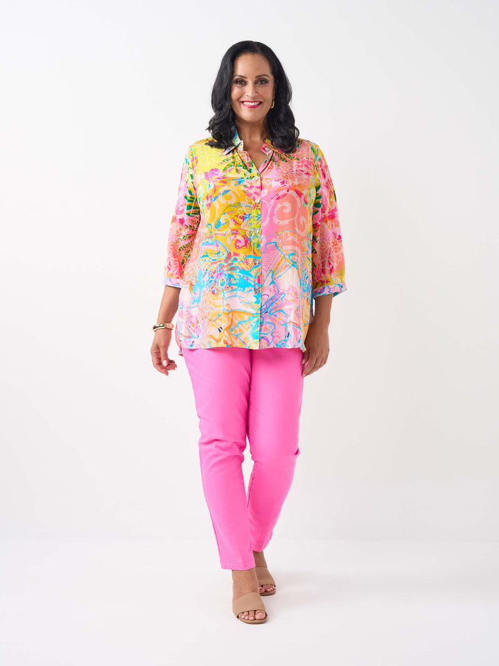 A woman waring a vibrant silky collared shirt with button front and colourful print in pink and yellow tone, paired with bright pink jeans