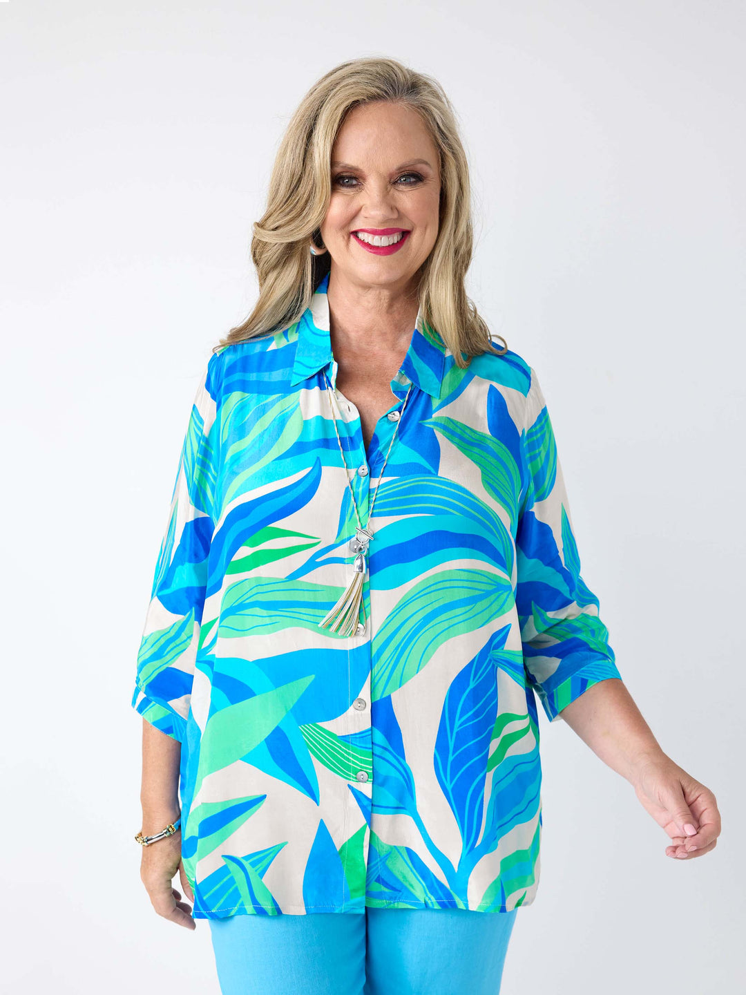 A woman wearing a silky collared shirt with aqua blue and green tropical leaves print, paired with aqua blue linen pant