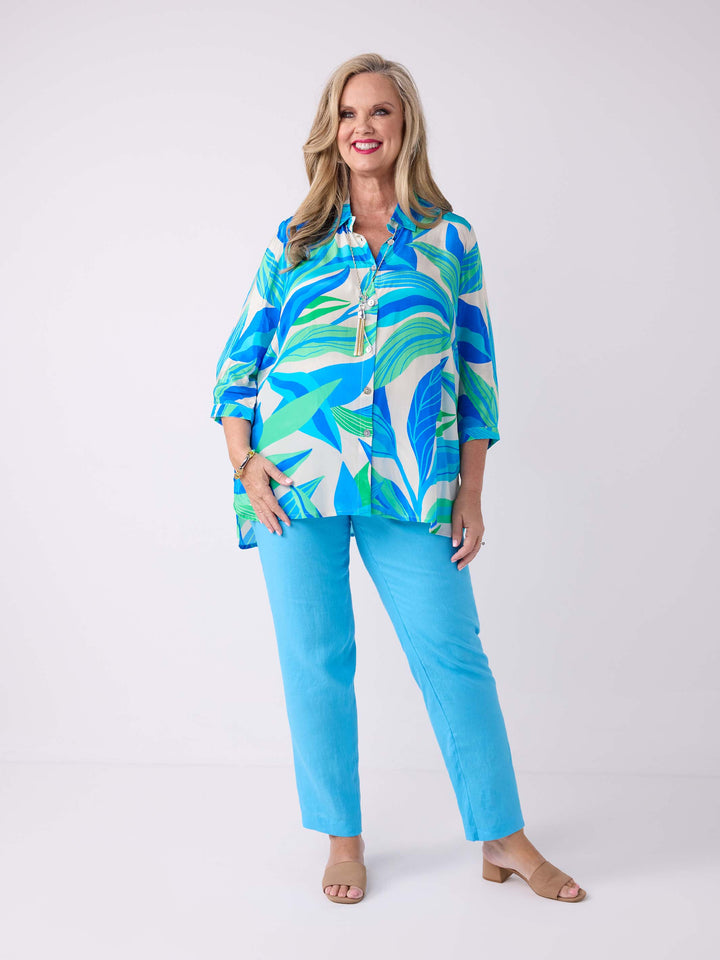 A woman wearing a silky collared shirt with aqua blue and green tropical leaves print, paired with aqua blue linen pant