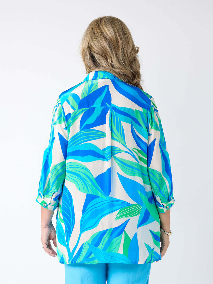 A woman viewed from the back wearing a silky collared shirt with aqua blue and green tropical leaves print.
