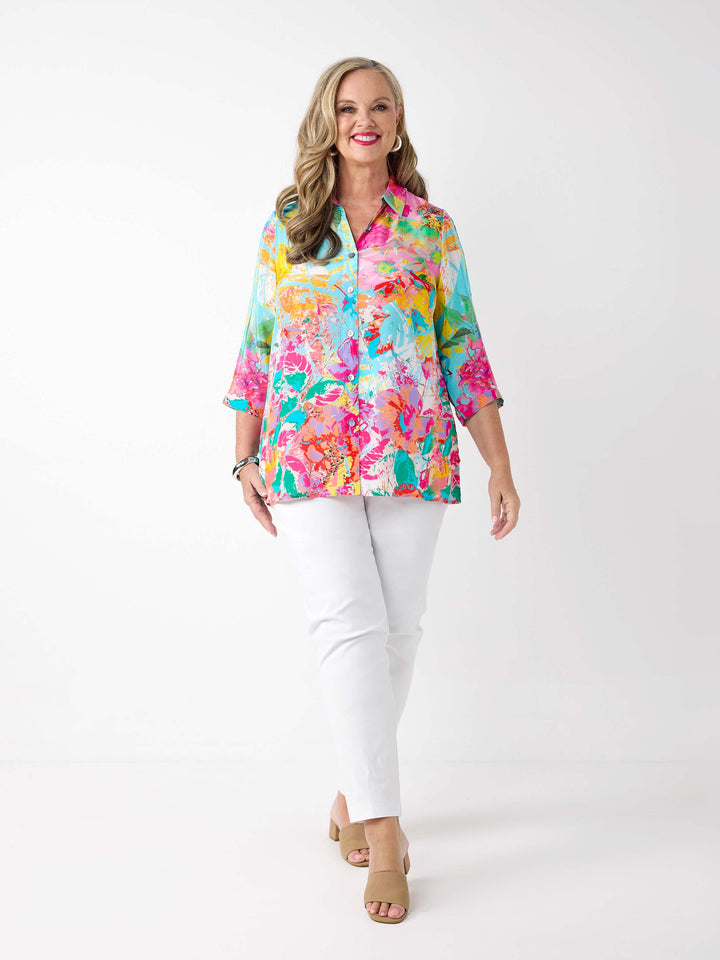 A woman wearing a vibrant abstract floral printed shirt in aqua blue and pink, paired with a pair of white pants