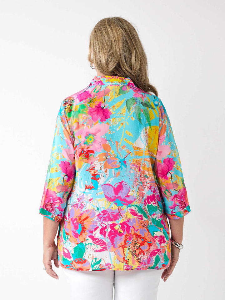 A woman wearing a vibrant abstract floral printed shirt in aqua blue and pink, paired with a pair of white pants