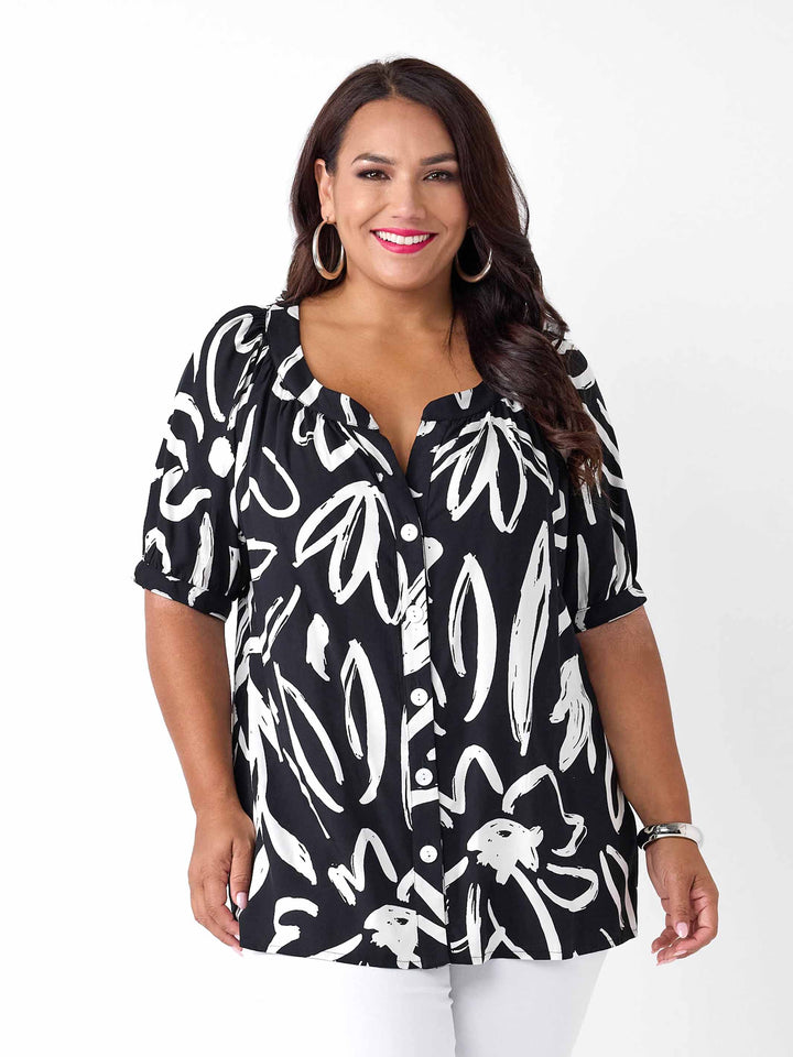 A woman wearing a black short-sleeve button-up blouse with a white abstract brushstroke print, paired with white pants