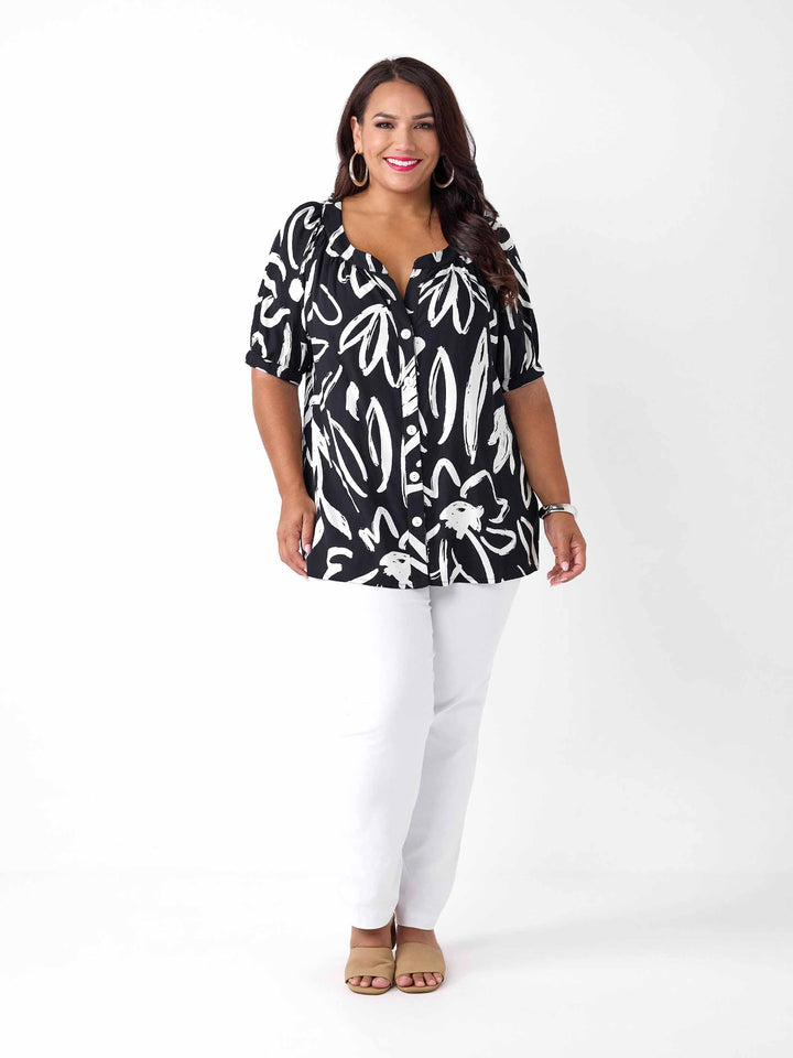 A woman wearing a black short-sleeve button-up blouse with a white abstract brushstroke print, paired with white pants