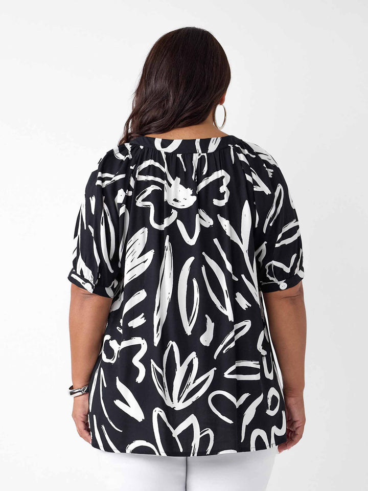 back view of a woman wearing a black short-sleeve button-up blouse with a white abstract brushstroke print, paired with white pants