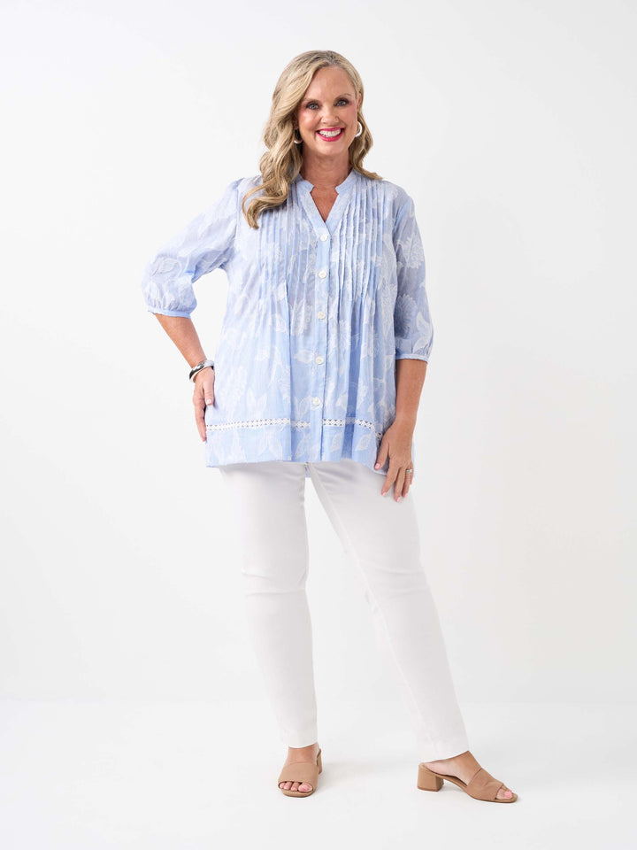 A  woman wearing a light blue and white patterned button-up blouse with pintuck detailing and white pants