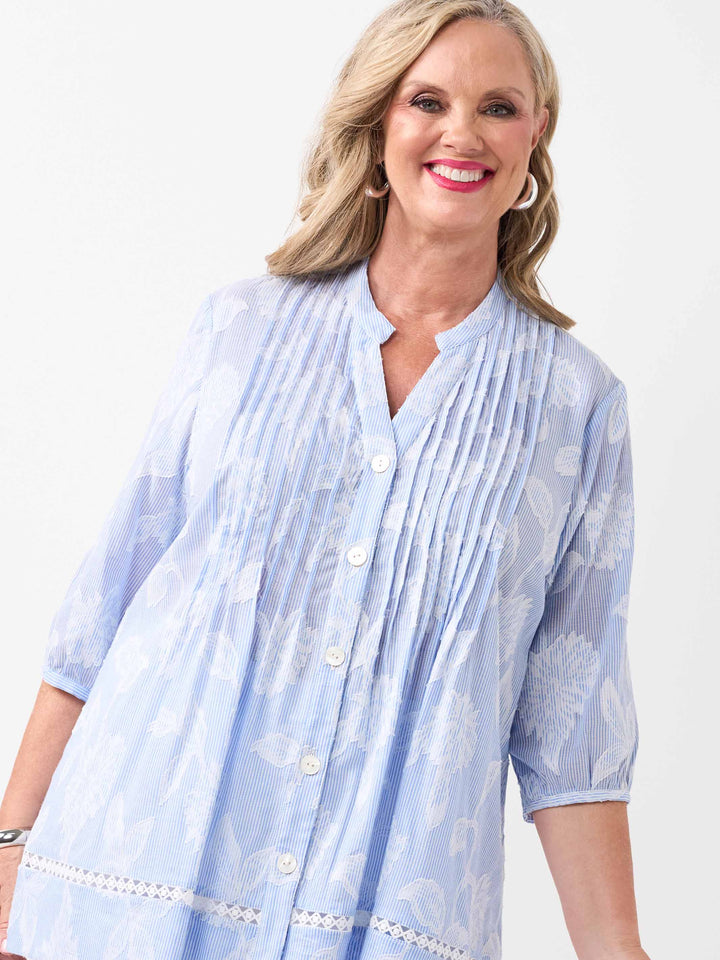 A woman wearing a light blue shirt with  white floral flocked design on a ticking stripe, pintuck front detailing, and white buttons