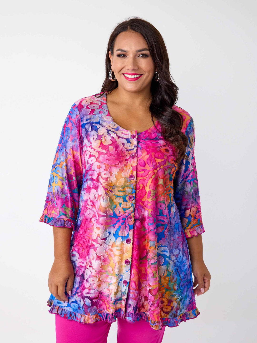 A woman wearing a vibrant, multicoloured floral burnout print tunic top with button through front and ruffle trim, paired with bright pink pants