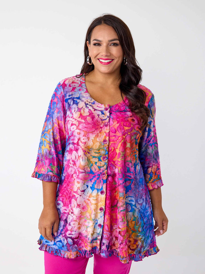 A woman wearing a vibrant, multicoloured floral burnout print tunic top with button through front and ruffle trim, paired with bright pink pants