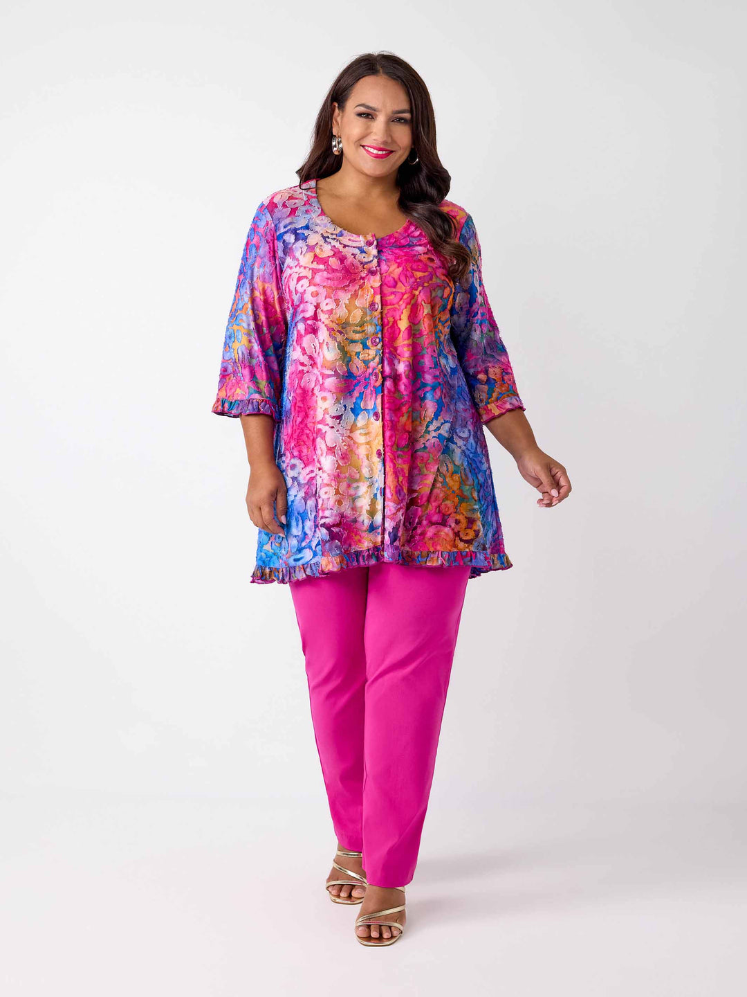 A woman wearing a vibrant, multicoloured floral burnout print tunic top with button through front and ruffle trim, paired with bright pink pants