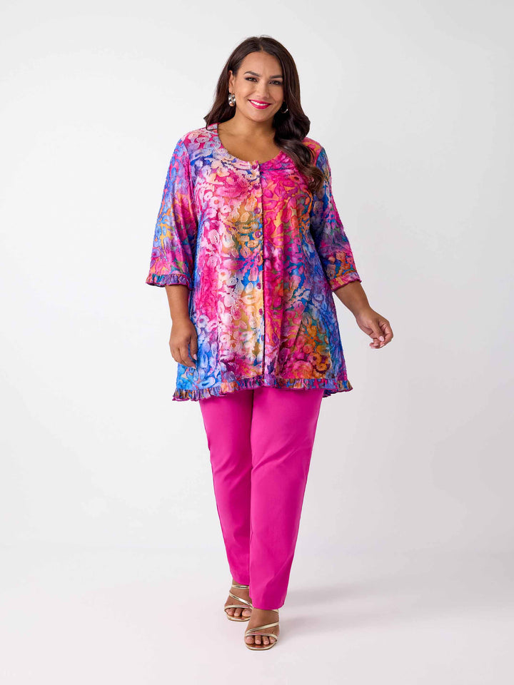 A woman wearing a vibrant, multicoloured floral burnout print tunic top with button through front and ruffle trim, paired with bright pink pants