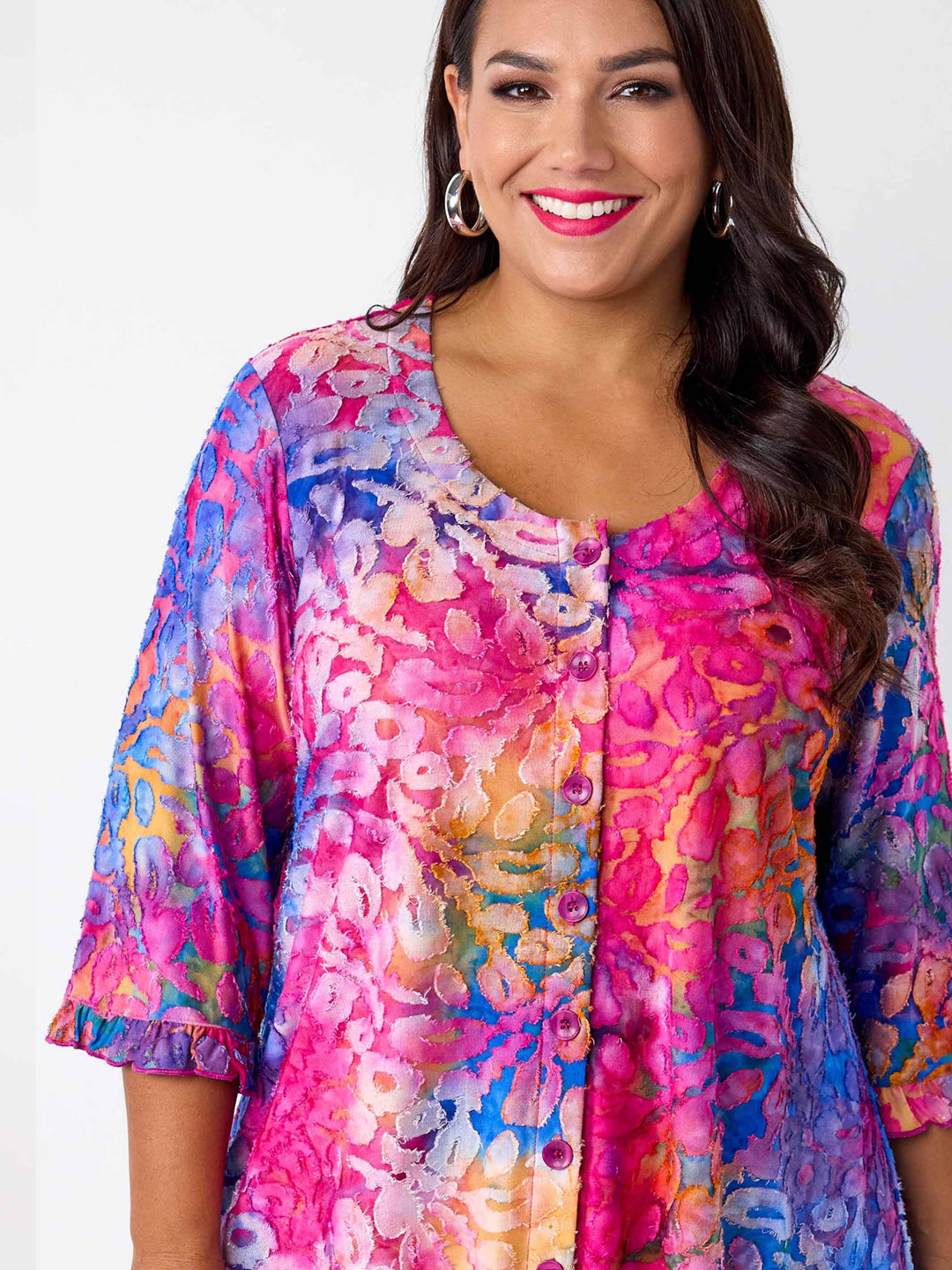 A woman wearing a vibrant, multicoloured floral burnout print tunic top with button through front and ruffle trim, paired with bright pink pants