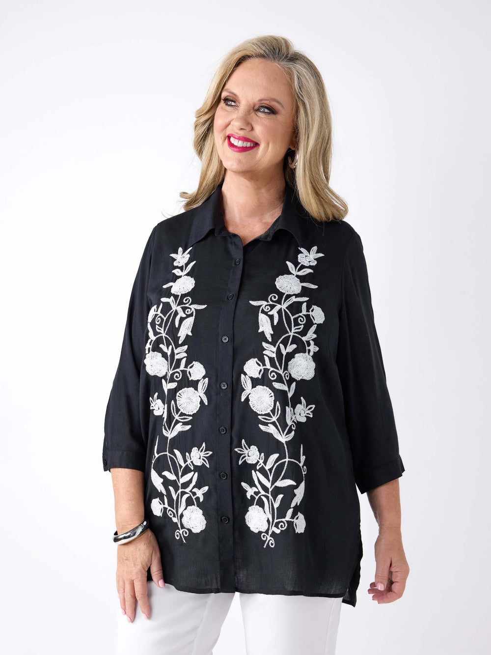 A woman wearing a black button-up shirt with white floral embroidery on the front, paired with white pants.