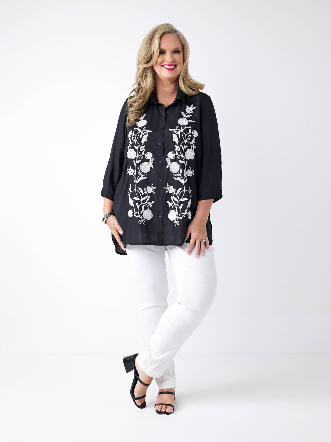 A woman wearing a black button-up shirt with white floral embroidery on the front, paired with white pants.