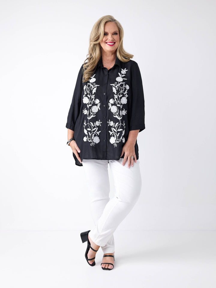A woman wearing a black button-up shirt with white floral embroidery on the front, paired with white pants.