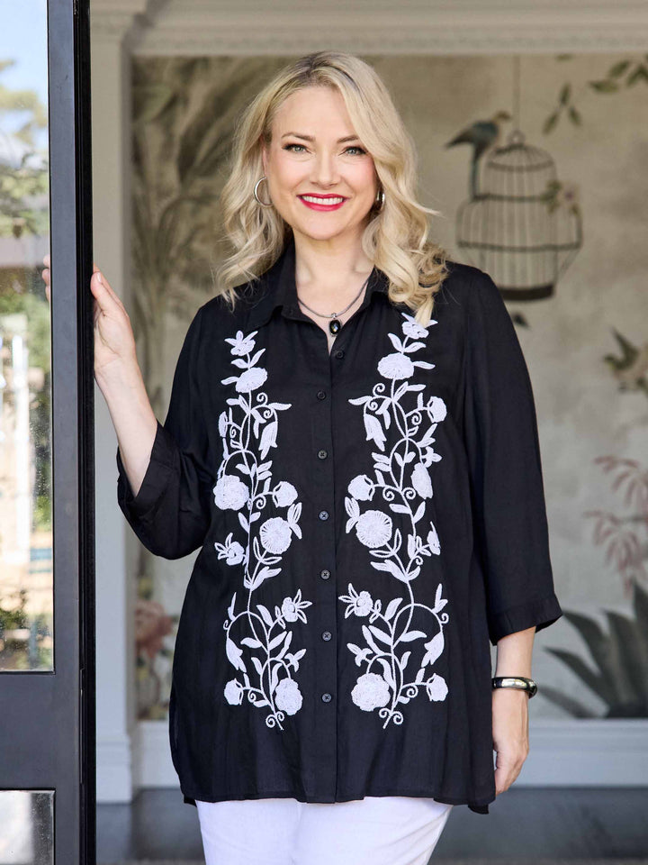 A woman wearing a black button-up shirt with white floral embroidery on the front, paired with white pants.