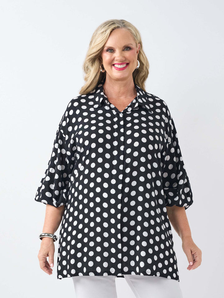 A woman wearing a black collared relaxed fit shirt with a white polka dot pattern, paired with white pants