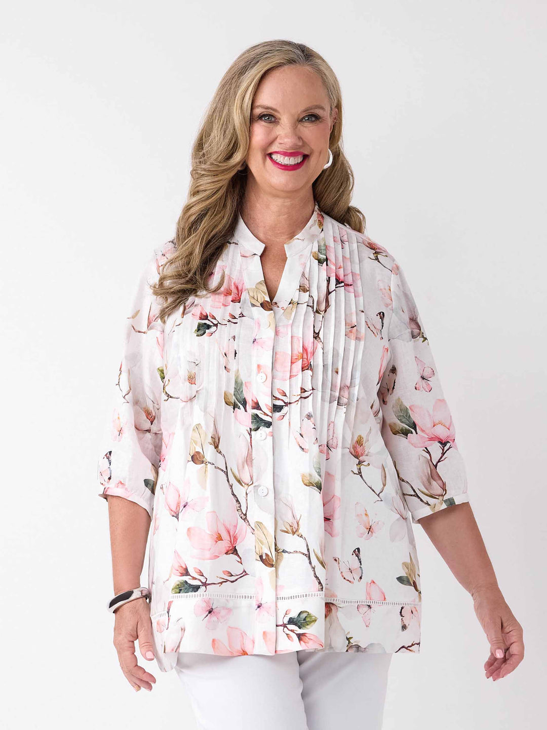 A woman wearing a button through pin tuck front top with pink magnolia print on white, paired with a pair of white pant