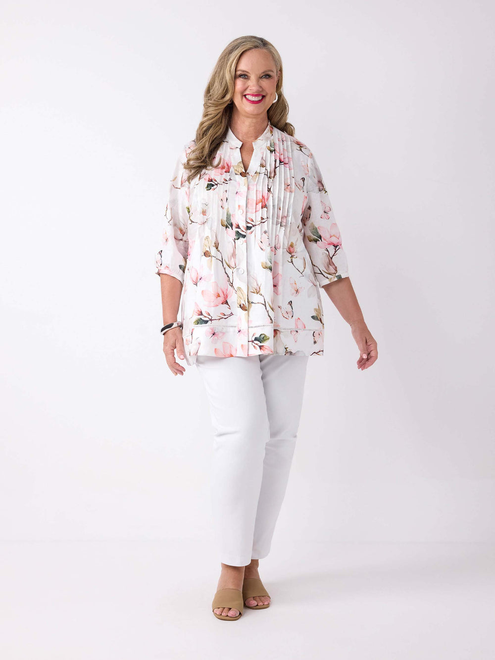 A woman wearing a button through pin tuck front top with pink magnolia print on white, paired with a pair of white pant