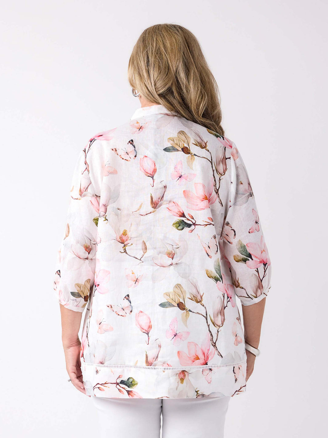 A woman wearing a button through pin tuck front top with pink magnolia print on white, paired with a pair of white pant