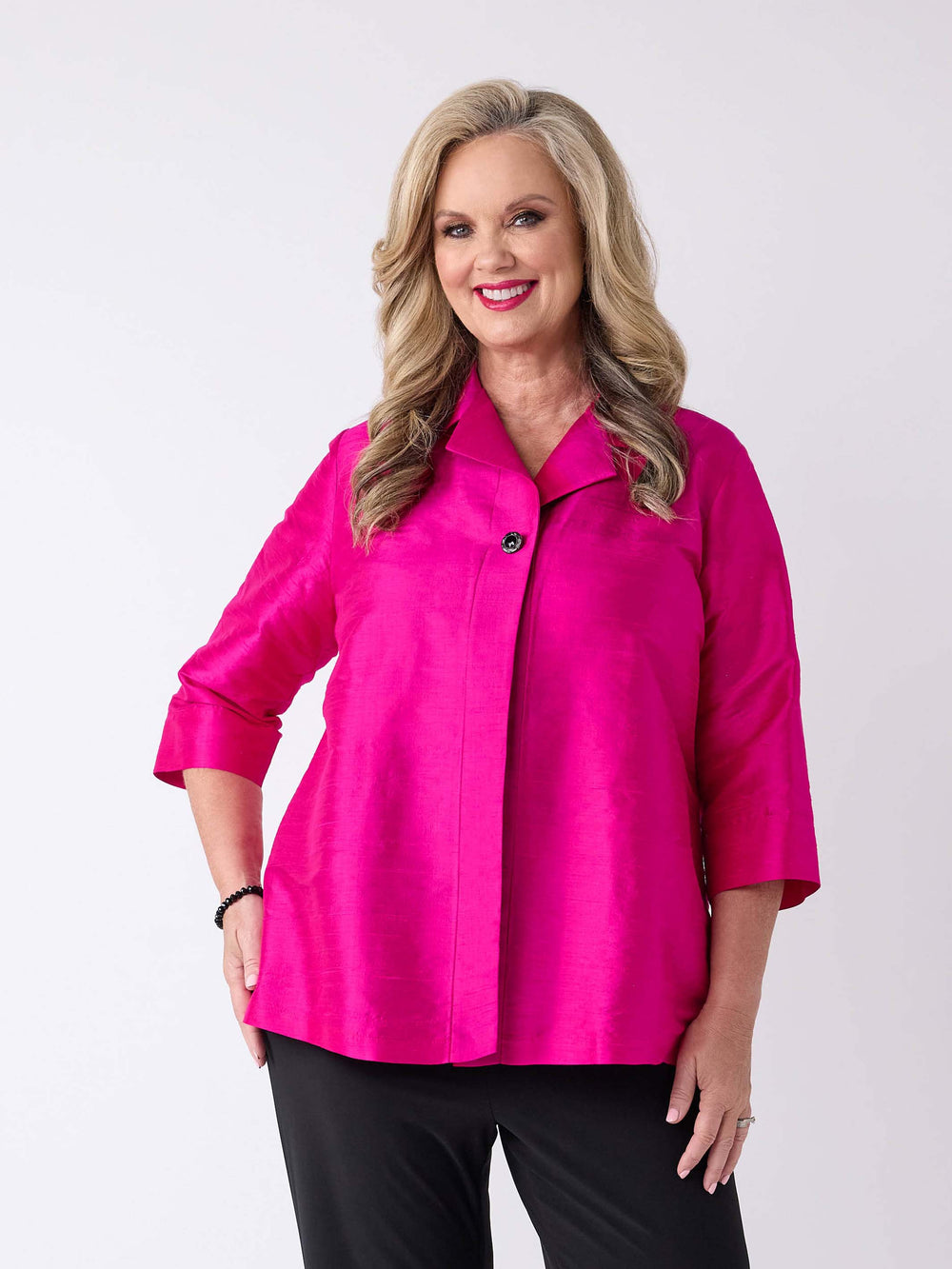 A woman wearing a v-neck collared silk shirt jacket in vibrant fuchsia pink with three-quarter sleeves and one-button front closure, paired with a pair of black pant