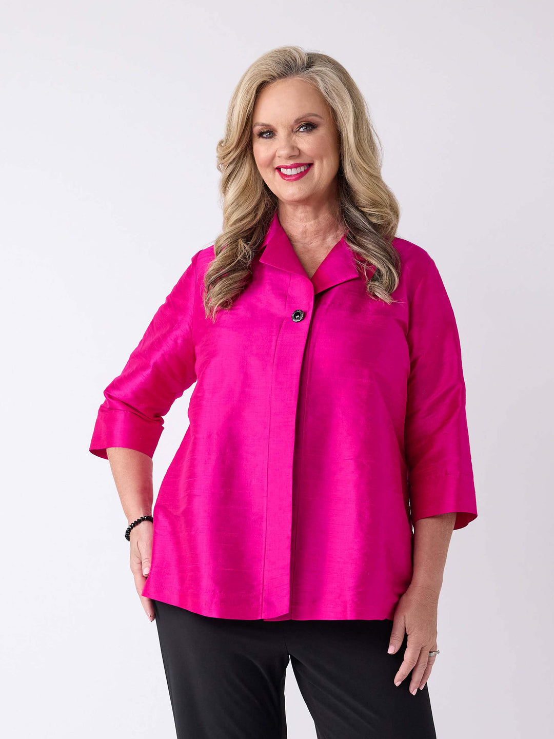A woman wearing a v-neck collared silk shirt jacket in vibrant fuchsia pink with three-quarter sleeves and one-button front closure, paired with a pair of black pant