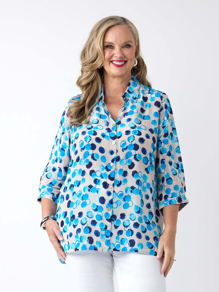 A woman wearing a light white button-up collared shirt with a vibrant blue and navy abstract watercolour dot pattern, paired with white pants