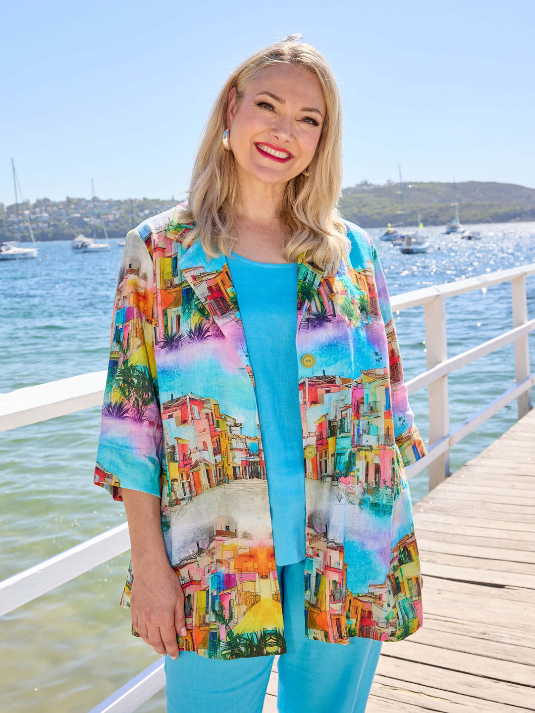 A woman wearing a bright multicolour printed collared linen jacket over a turquoise linen cami and matching pants. 
