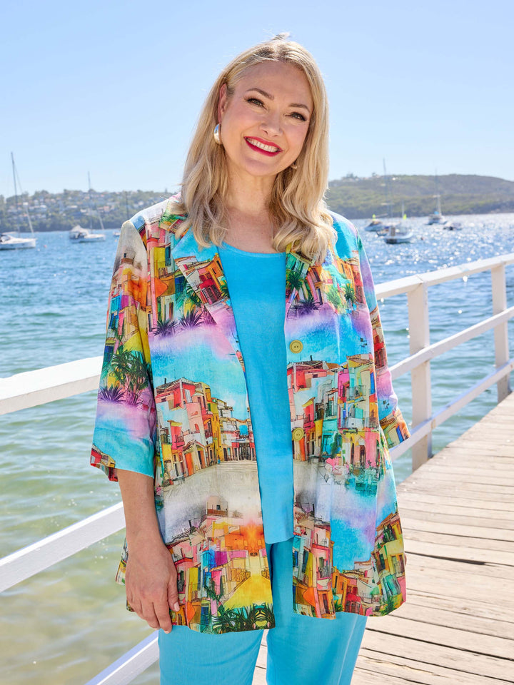 A woman wearing a bright multicolour printed collared linen jacket over a turquoise linen cami and matching pants. 