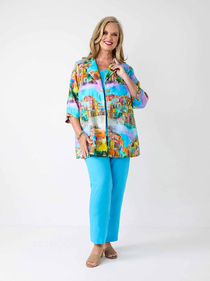 A woman wearing a colourful short-sleeve collared linen jacket with a vibrant tropical beach print, paired with bright turquoise pants